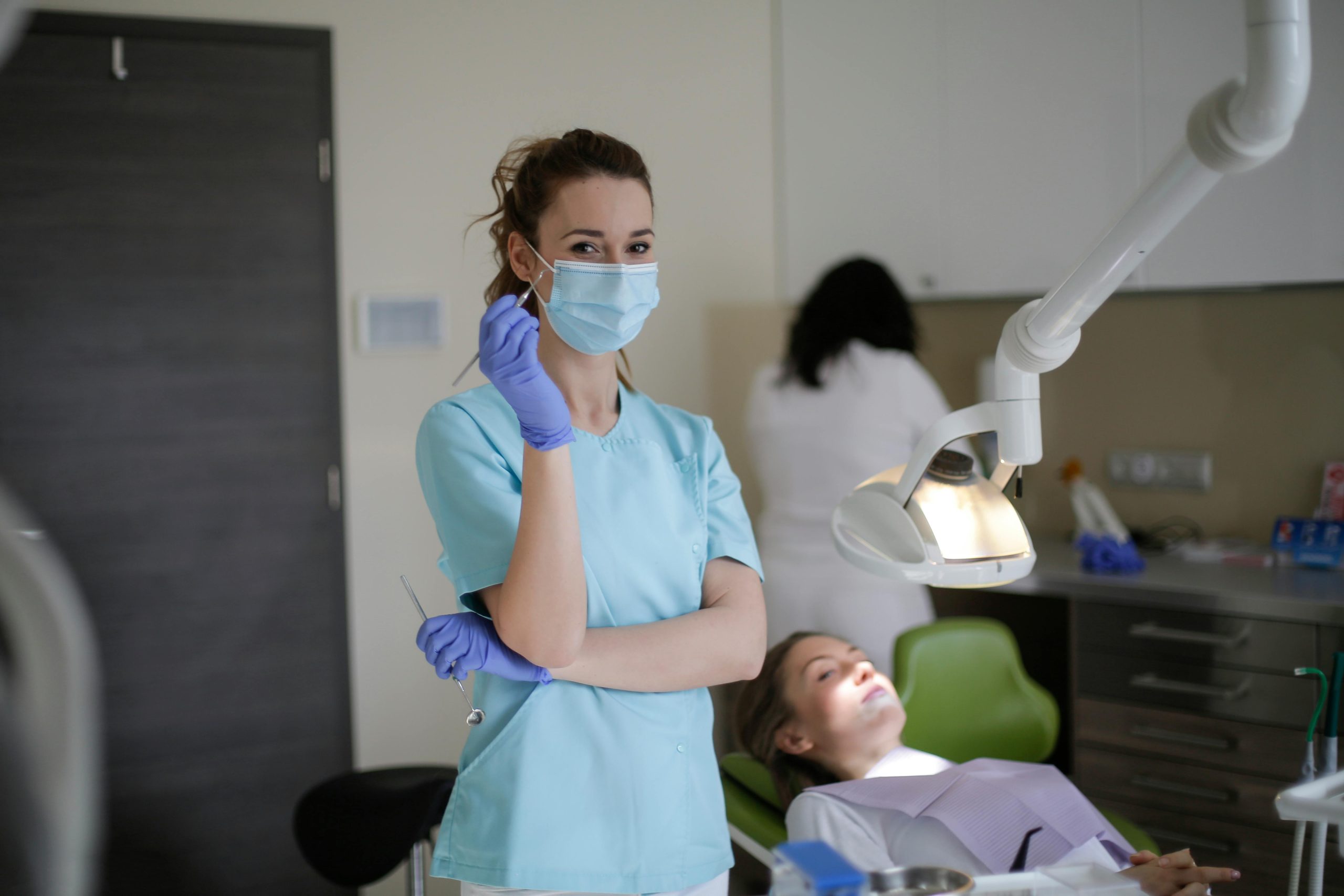 professional-dental-checkup-clinic A female dentist in blue scrubs and a mask holding tools, with a patient resting in a dental chair in the background